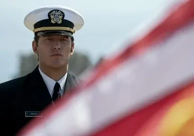 Homem sério em uniforme da marinha, com boné branco, olhando fixamente. Bandeira americana borrada no primeiro plano.
