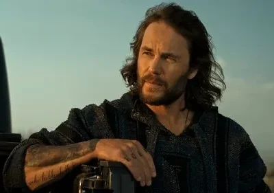 Homem barbudo de cabelos longos, camisa escura, braço tatuado. Encosta em caminhonete, olhando pensativo para o deserto azulado.