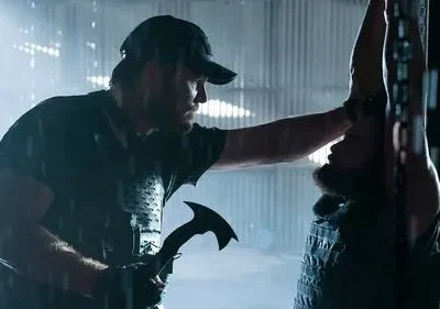 Homem de boné com machado encara outro barbudo pendurado de cabeça para baixo em correntes. Cenário escuro e tenso.