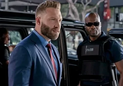 Homem barbudo de terno azul e segurança de colete tático e óculos escuros. Cena urbana com carro e clima sério.