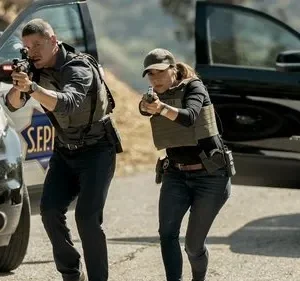 Um homem e uma mulher em coletes táticos e armados avançam, usando carros como cobertura. Um terceiro agente também armado no fundo. Ação intensa.