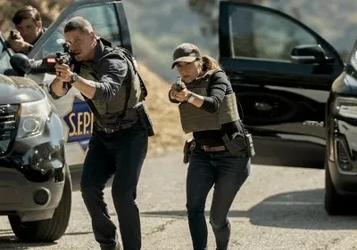 Um homem e uma mulher em coletes táticos e armados avançam, usando carros como cobertura. Um terceiro agente também armado no fundo. Ação intensa.