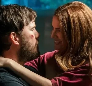 Mulher de cabelos ruivos sorri ao abraçar homem barbudo com ferimento no rosto. Olham-se com ternura num ambiente escuro.