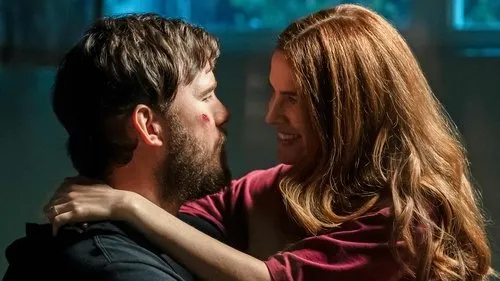 A Lista Terminal – T01E07:Extinção 1 Mulher de cabelos ruivos sorri ao abraçar homem barbudo com ferimento no rosto. Olham-se com ternura num ambiente escuro.