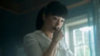 Mulher de franja, camisa branca, com a mão na boca, olha pensativa em um quarto escuro com janela iluminada. Clima misterioso.