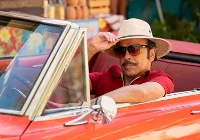 Homem de bigode, óculos de sol e chapéu em um conversível vermelho vibrante. Ele segura o chapéu com ar confiante.