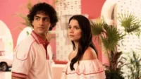 Um jovem de polo rosa e uma jovem de blusa ombro a ombro rosa. Ambiente retrô tropical, paredes rosas e plantas. Clima vibrante.