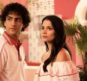 Um jovem de polo rosa e uma jovem de blusa ombro a ombro rosa. Ambiente retrô tropical, paredes rosas e plantas. Clima vibrante.