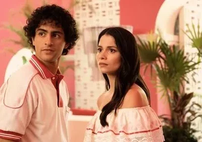 Um jovem de polo rosa e uma jovem de blusa ombro a ombro rosa. Ambiente retrô tropical, paredes rosas e plantas. Clima vibrante.