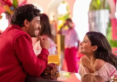 Um homem de camisa vermelha sorri para uma mulher de blusa rosa em um ambiente vibrante com arcos e plantas, transmitindo romance.