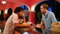 Dois homens em balcão de hotel retrô. Um fala ao telefone, outro loiro tem expressão surpresa. Ambiente rosa, vibe anos 80.