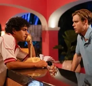 Dois homens em balcão de hotel retrô. Um fala ao telefone, outro loiro tem expressão surpresa. Ambiente rosa, vibe anos 80.