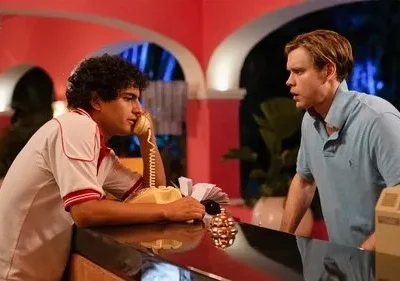 Dois homens em balcão de hotel retrô. Um fala ao telefone, outro loiro tem expressão surpresa. Ambiente rosa, vibe anos 80.