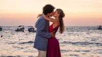 Casal se beija na praia ao pôr do sol. Ele de blazer claro, ela de vestido vermelho/roxo. Barcos no mar. Cena romântica.