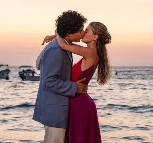 Casal se beija na praia ao pôr do sol. Ele de blazer claro, ela de vestido vermelho/roxo. Barcos no mar. Cena romântica.
