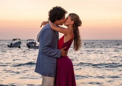 Casal se beija na praia ao pôr do sol. Ele de blazer claro, ela de vestido vermelho/roxo. Barcos no mar. Cena romântica.