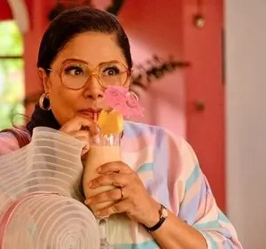 Mulher com óculos e expressão divertida bebe coquetel tropical em cenário vibrante rosa. Veste blusa listrada pastel.