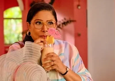 Mulher com óculos e expressão divertida bebe coquetel tropical em cenário vibrante rosa. Veste blusa listrada pastel.