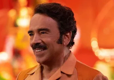 Homem sorridente de bigode e camisa laranja, em fundo vibrante de tons quentes e luzes borradas. Clima festivo.