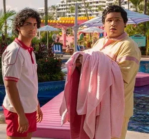 Dois rapazes em uniforme de resort. Um sério, outro surpreso com toalhas rosa, em piscina vibrante e tropical.