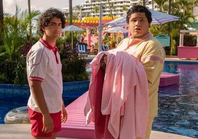 Dois rapazes em uniforme de resort. Um sério, outro surpreso com toalhas rosa, em piscina vibrante e tropical.