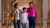 Uma família feliz (mulher, menino, homem) caminha sorrindo em um interior iluminado. O homem usa camisa rosa vibrante.