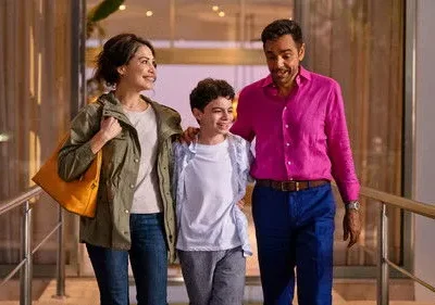 Uma família feliz (mulher, menino, homem) caminha sorrindo em um interior iluminado. O homem usa camisa rosa vibrante.