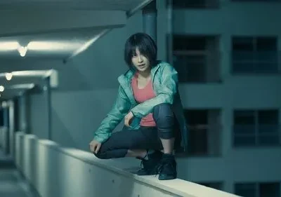 Mulher jovem de jaqueta verde-água agachada em muro alto. Fundo urbano noturno com luzes. Olhar atento e clima tenso.