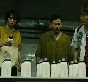 Quatro personagens (3 homens, 1 mulher) com expressões tensas olham garrafas em sala industrial escura. Clima de desafio.