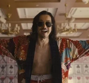 Homem de óculos escuros, cabelo comprido e roupão colorido, braços abertos e rindo, em sala luxuosa com símbolos de cartas.