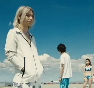 Três jovens num telhado sob céu azul. Uma loira em hoodie à frente, outros dois em trajes de verão. Clima vasto e pensativo.