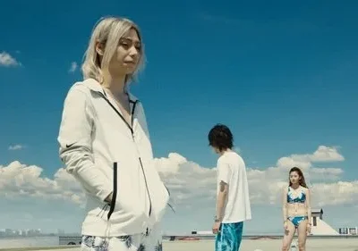 Três jovens num telhado sob céu azul. Uma loira em hoodie à frente, outros dois em trajes de verão. Clima vasto e pensativo.