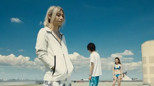Três jovens num telhado sob céu azul. Uma loira em hoodie à frente,outros dois em trajes de verão. Clima vasto e pensativo.