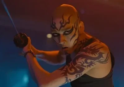Pessoa careca com pintura facial e tatuagens, empunhando uma katana em pose de combate, sob luzes neon dramáticas.