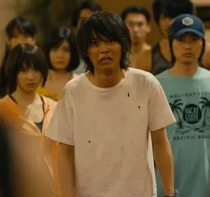 Um grupo de jovens assustados em ambiente fechado. Um homem de camisa branca suja com expressão de pânico se destaca no centro.