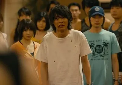 Um grupo de jovens assustados em ambiente fechado. Um homem de camisa branca suja com expressão de pânico se destaca no centro.