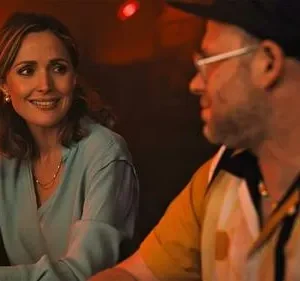 Mulher sorridente de blusa azul em bar com luz avermelhada, olhando para homem de boné e óculos. Clima aconchegante.