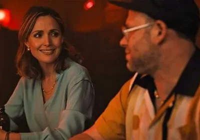 Mulher sorridente de blusa azul em bar com luz avermelhada, olhando para homem de boné e óculos. Clima aconchegante.