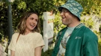Uma mulher loira sorri com um homem barbudo de chapéu e jaqueta verde. Eles parecem felizes ao ar livre, sob o sol.