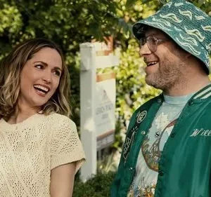 Uma mulher loira sorri com um homem barbudo de chapéu e jaqueta verde. Eles parecem felizes ao ar livre, sob o sol.