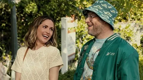 Uma mulher loira sorri com um homem barbudo de chapéu e jaqueta verde. Eles parecem felizes ao ar livre,sob o sol.