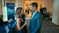 Um homem de terno azul e uma mulher de vestido preto sorriem enquanto conversam em um evento profissional com banner Partners' Retreat.