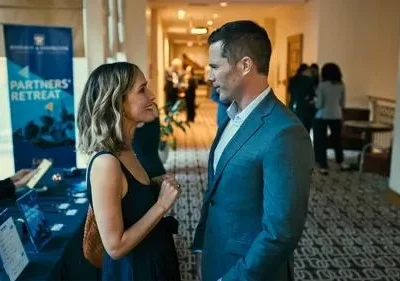 Um homem de terno azul e uma mulher de vestido preto sorriem enquanto conversam em um evento profissional com banner Partners' Retreat.