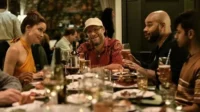 Jantar animado com quatro pessoas: mulher de laranja, homem de boné e barba sorrindo, e dois homens conversando em uma mesa.