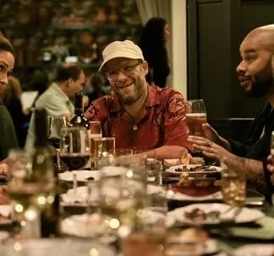 Jantar animado com quatro pessoas: mulher de laranja, homem de boné e barba sorrindo, e dois homens conversando em uma mesa.