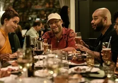 Jantar animado com quatro pessoas: mulher de laranja, homem de boné e barba sorrindo, e dois homens conversando em uma mesa.