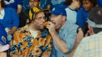 Dois homens comendo um cachorro-quente num estádio. Um usa camisa de tigre e chapéu, o outro um boné. Clima divertido e casual.