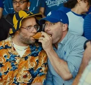 Dois homens comendo um cachorro-quente num estádio. Um usa camisa de tigre e chapéu, o outro um boné. Clima divertido e casual.