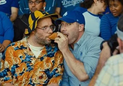 Dois homens comendo um cachorro-quente num estádio. Um usa camisa de tigre e chapéu, o outro um boné. Clima divertido e casual.