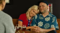 Casal sorridente num bar. Mulher loira encosta no ombro de homem barbudo de camisa florida. Bebem cervejas, clima afetuoso.